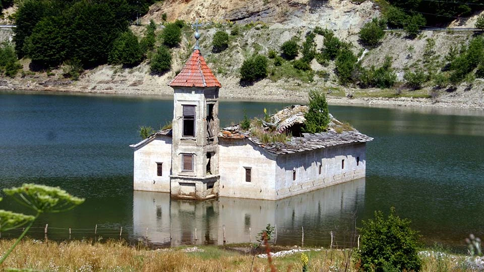 This image shows an old, partially ruined stone church sitting in the middle of a lake, with water reaching almost halfway up its walls.  The building is rectangular, made of light-colored stone, and has a small tower with a red-tiled roof rising from one side. The tower looks weathered, with missing windows and broken edges. The rest of the church’s roof has collapsed, leaving wooden beams exposed and small plants growing on top, as if nature has slowly taken over.  The lake around the church is calm and greenish-blue, reflecting parts of the building. In the background, steep hills rise up, covered with patches of grass, shrubs, and scattered trees. The scene feels quiet and abandoned — a lonely structure standing in the water, hinting at history, time, and the power of nature reclaiming old places.