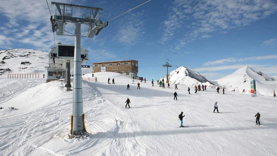 This image shows a lively winter scene at a snowy mountain ski resort. The ground is completely covered in bright white snow, and many people are skiing or snowboarding down a gentle slope. They are spread out across the scene, moving in different directions, creating a sense of activity and motion.  In the center stands a tall ski-lift pole made of metal, supporting thick cables that run across the image. Attached to the cables are ski-lift chairs used to carry people up the mountain, though none of the chairs are visible up close.  Behind the pole, there is a modern ski-lift station — a structure with large windows and machinery that powers the lift. Nearby is a simple rectangular building, wooden or beige in color, sitting on a small rise of snow.  The background shows beautiful snow-covered mountain peaks under a bright blue sky with scattered thin clouds. The whole scene feels open, crisp, and full of winter energy, with skiers enjoying the clear weather and fresh snow at a high-altitude ski area.